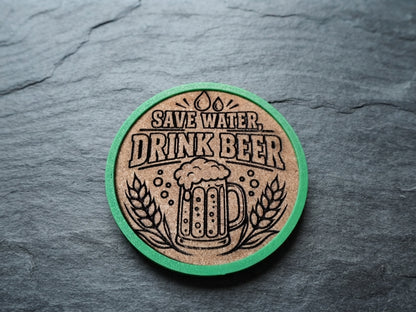 A round cork coaster with a green rim, featuring a black engraved design of a beer mug, wheat stalks, and the text 'SAVE WATER, DRINK BEER.