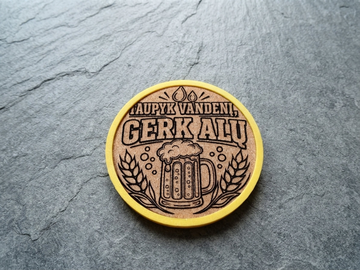 A round wooden coaster with a yellow border, featuring a laser-engraved beer mug and wheat design with the Lithuanian text 'TAUPYK VANDENI, GERK ALU', placed on a dark gray slate surface.