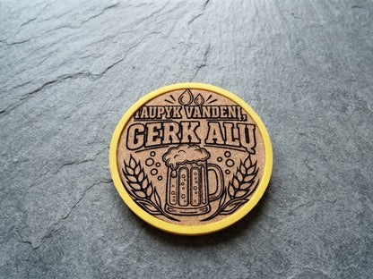 A round wooden coaster with a yellow border, featuring a laser-engraved beer mug and wheat design with the Lithuanian text 'TAUPYK VANDENI, GERK ALU', placed on a dark gray slate surface.