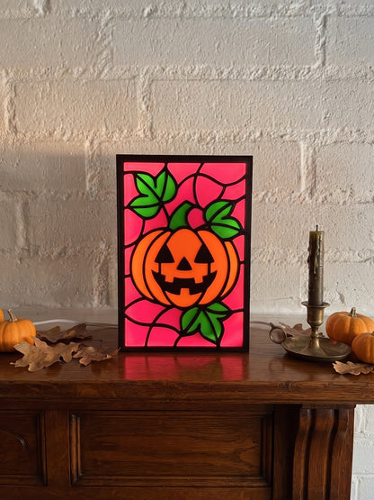 A framed stained-glass-style pumpkin artwork with a carved orange pumpkin on a magenta background, displayed on a wooden table against a white brick wall with small pumpkins and a candle nearby.