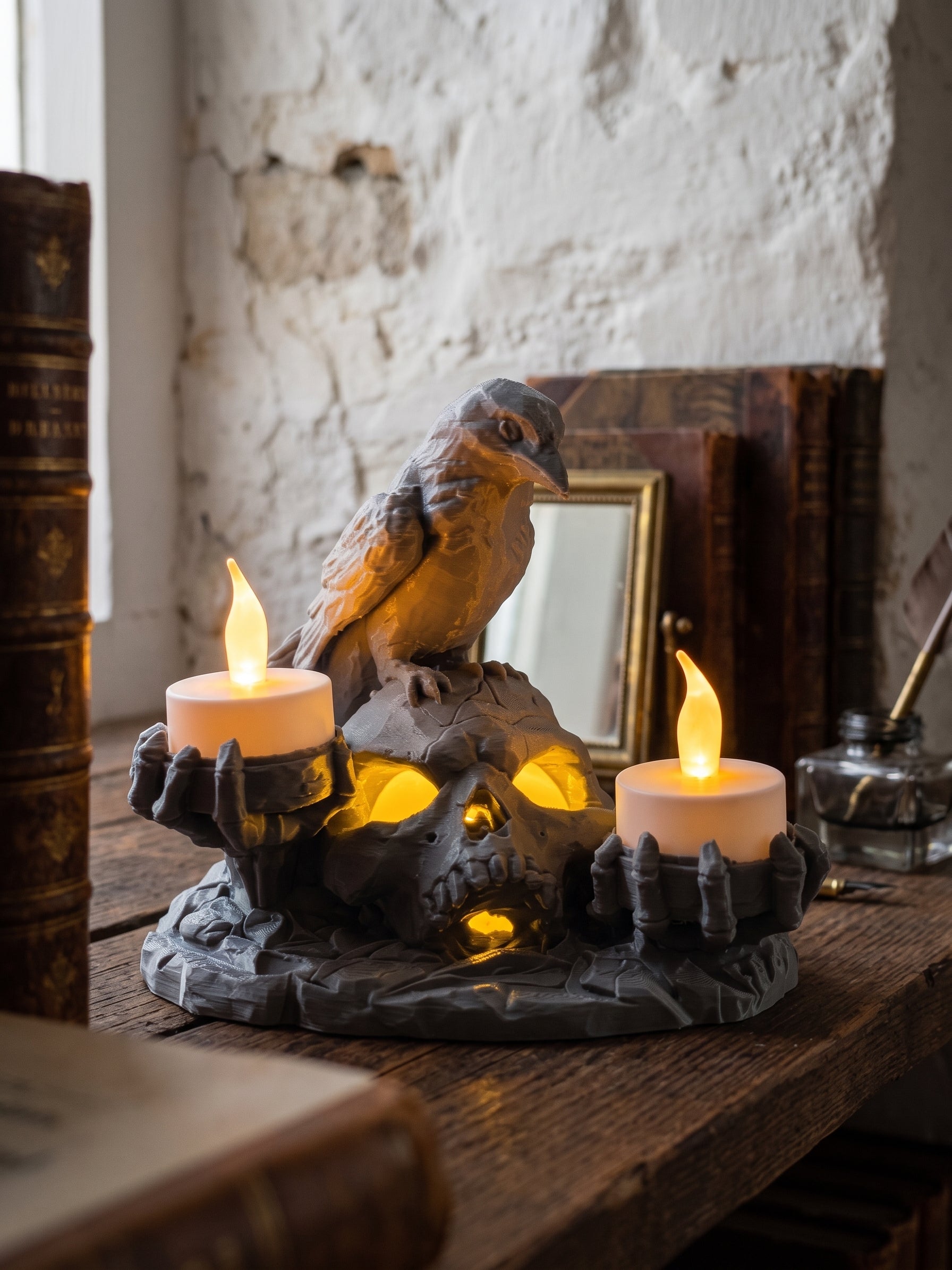 A gothic-style skull candle holder with a raven perched on top, holding two lit white pillar candles, displayed on a rustic wooden table in a vintage room setting.