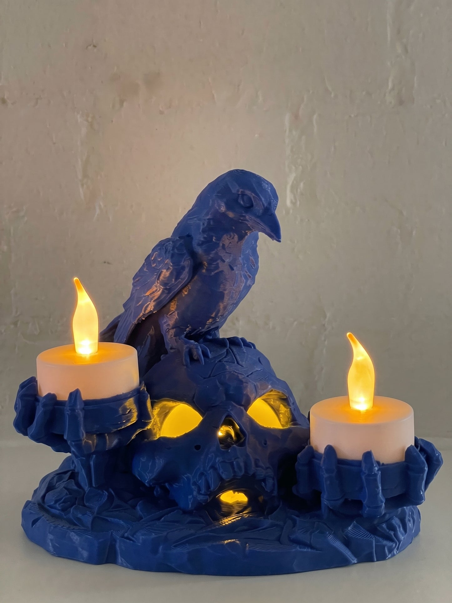 A blue raven figurine perched on a skull-shaped holder with three lit white candles, set against a textured off-white wall.